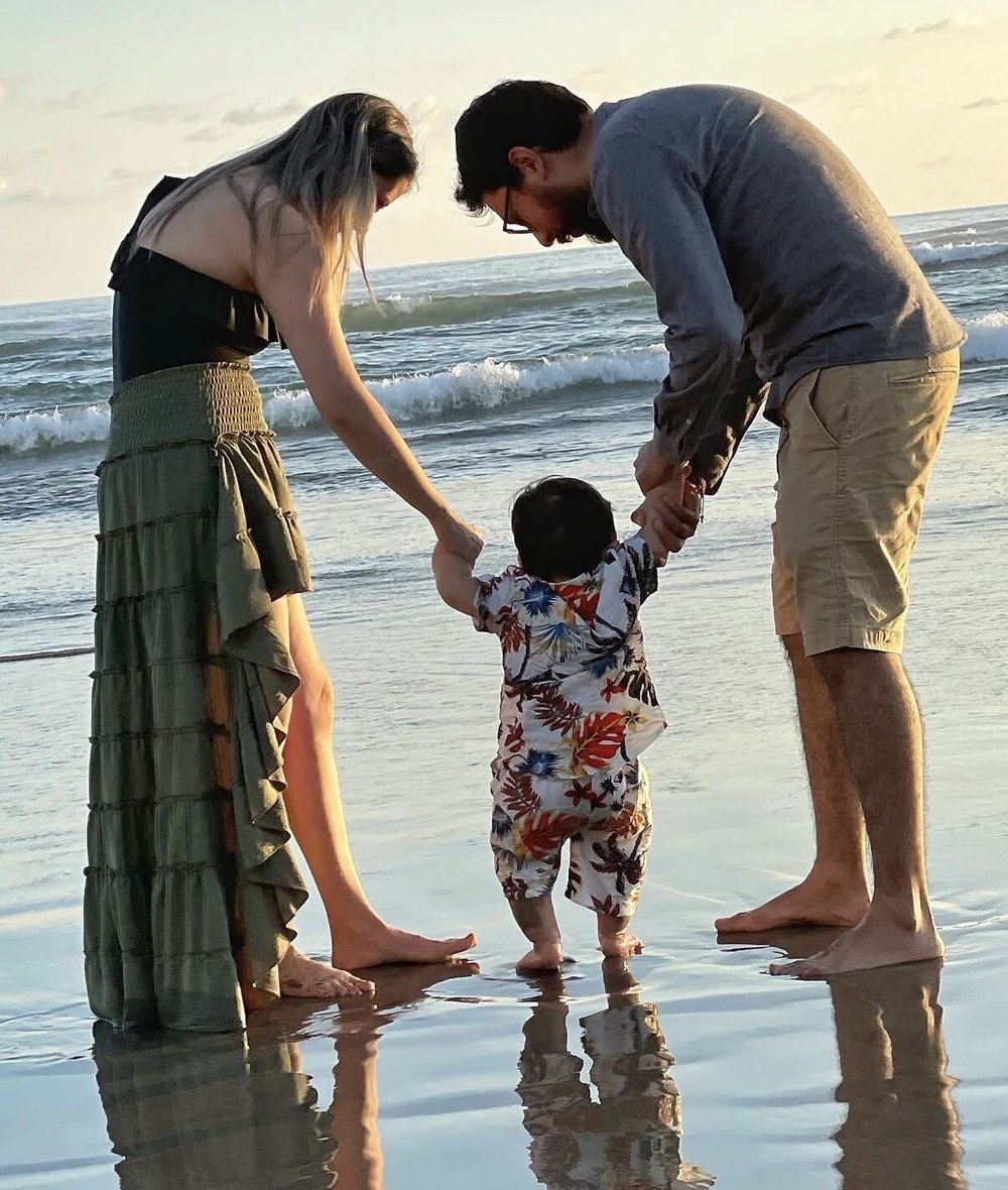 Una mamá y un papá se inclinan hacia su bebé, cada uno de una mano, mientras el niño da pasos sobre la arena mojada al atardecer.