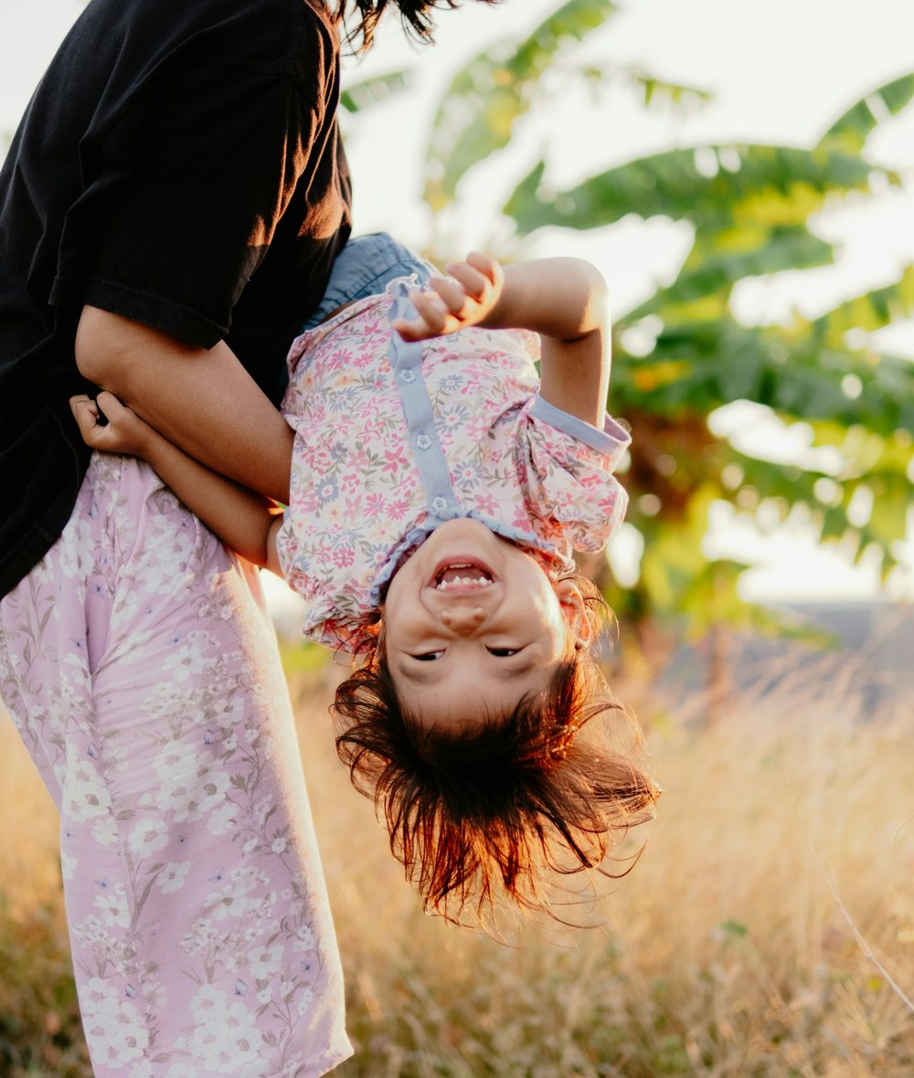 Una mamá carga de cabeza a su hija pequeña, que se ríe, en un campo dorado a contraluz.